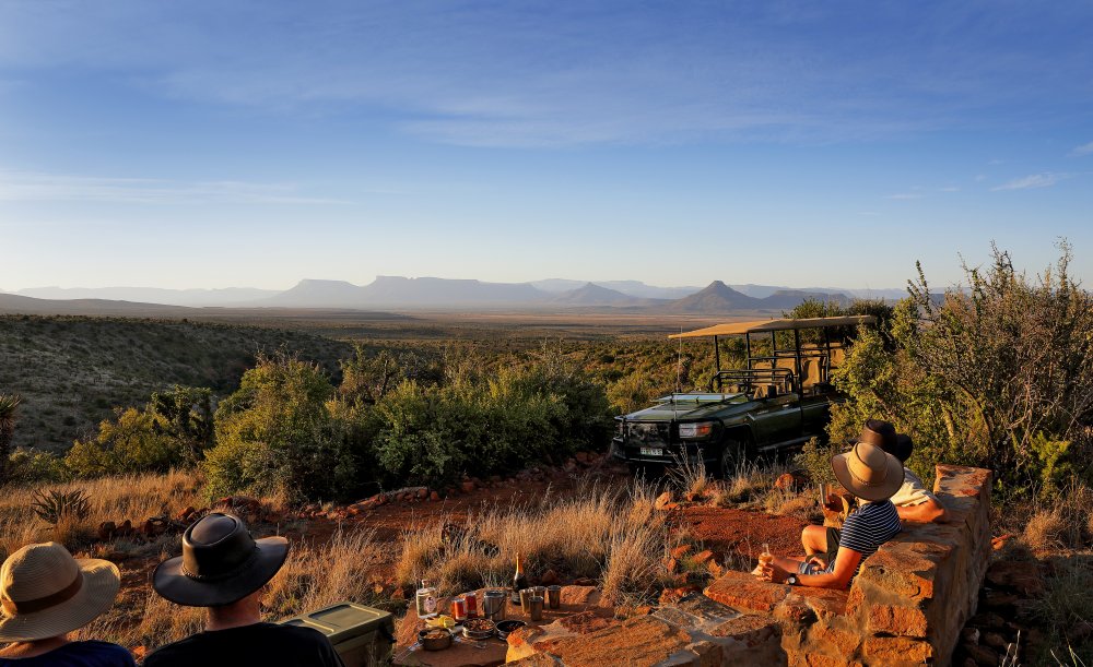 Safari travelers enjoy sunset view beside an open vehicle.